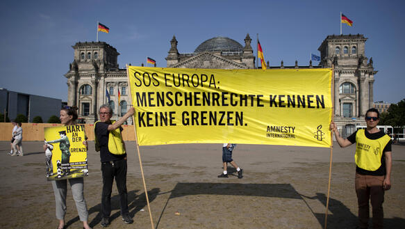 Vier Menschen vor dem Reichstagsgebäude, dem Sitz des Deutschen Bundestag. Zwei davon halten ein großes gelbes Amnesty-Transparent hoch, auf dem steht: SOS Europa – Menschenrecht kennen keine Grenzen. 