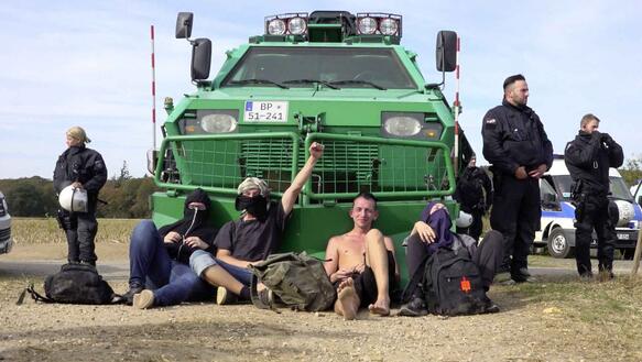 Das Foto zeigt ein großes grünes Fahrzeug der Bundespolizei und vier teilweise vermummte Menschen, die davor auf dem Boden sitzen. Eine Person hebt die linke Faust in den Himmel.