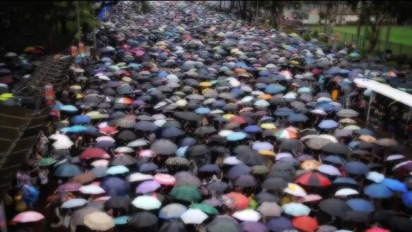 Viele Menschen auf einer Straße tragen Regenschirme, sodass sich ein Mosaik aus vielen hunderten Schirmen aus Vogelperspektive ergibt.