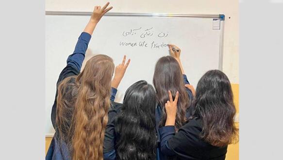 Das Foto zeigt vier Mädchen bzw. junge Frauen mit langen Haaren von hinten fotografiert. Drei von ihnen heben eine Hände und formen mit Zeige- und Mittelfinger das Victory-Zeichen. Eine von ihnen schreibt auf die Tafel.