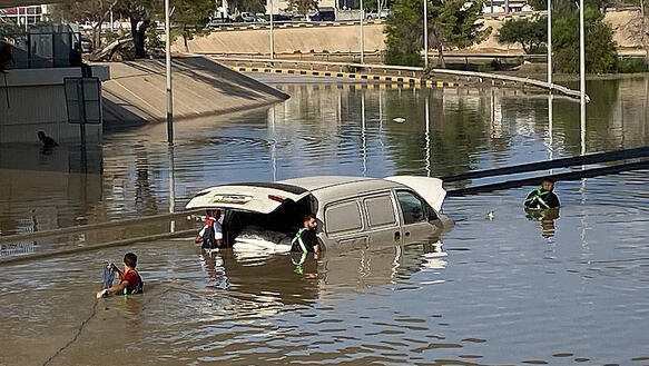 Das Foto zeigt eine komplett überflutete Straße bei Tag. Ein Kleintransporter ragt mit geöffneter Heckklappe zur Hälte aus dem Wasser heraus. Vier Personen waten durch das Wasser in der Nähe des Fahrzeugs.