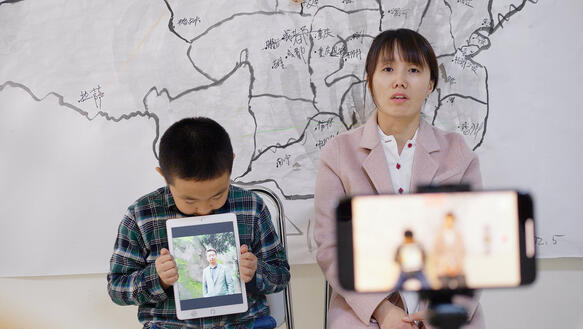 Das Bild zeigt ein Kind und eine Frau, die nebeneinander sitzen. Das Kind hält ein Tablet in der Hand, darauf zu sehen das Foto eines Mannes. Die Frau nimmt gleichzeitig ein Video von sich mit dem Smartphone auf.