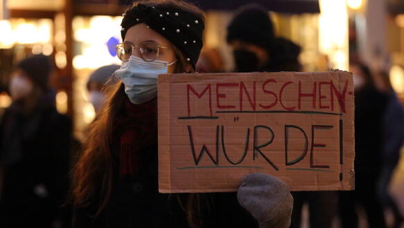 Das Bild zeigt eine jungeFrau mit einem Protestschild "Menschen Würde"