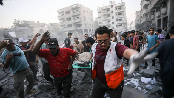 Das Foto zeigt Männer, die einen Mann auf einer Trage tragen, inmitten von Schutt und zerstörten Gebäuden.