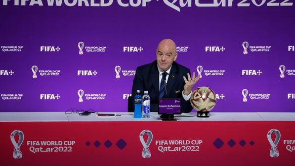 Das Bild zeigt eine Person, die hinter einem langem Tisch sitzt, dahinter ein Banner mit der Aufschrift "FIFA World CUp 2022"