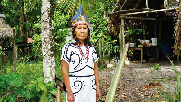 Eine indigene Frau mittleren Alters in tradtionellem Gewand, sie trägt eine Halskette und Federschmuck auf dem Kopf und steht im Wald vor einem Baum, im Hintergrund eine Hütte.