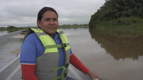 Ein Frau mit Rettungsweste sitzt in einem kleinen Boot und fährt an der Nähe des Ufers auf einem Fluss.