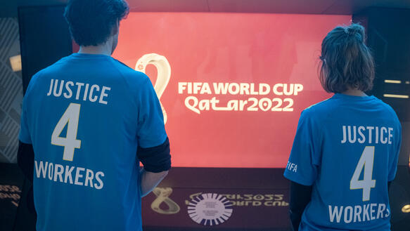 Das Bild zeigt zwei Personen von hinten, die auf einen Bildschirm schauen. Darauf steht "FIFA World Cup"