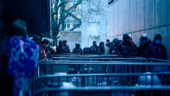 Das Bild zeigt Absperrgitter, mehrere Menschen stellen sich in einer Reihe an und warten