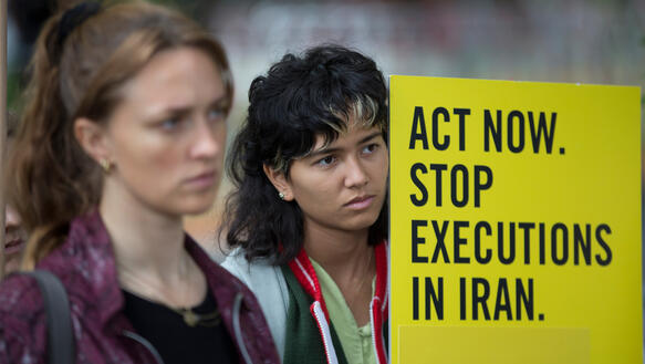 Das Bild zeigt zwei Frauen, die beide ernst schauen und von denen eine, ein gelbes Schild mit der schwarzen Aufschrift "Act now. Stop executions in Iran." hält.