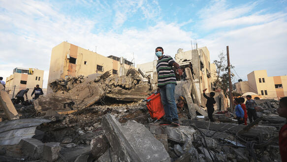 Männer suchen nach Überlebenden in einem zerbombten Haus, Kinder stehen daneben.