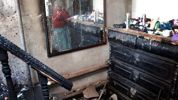 Das Bild zeigt den Blick in einen Hausflur, der durch einen Brand verwüstet ist. Durch einen Spiegel sieht man eine ältere Frau die auf den Boden schaut.