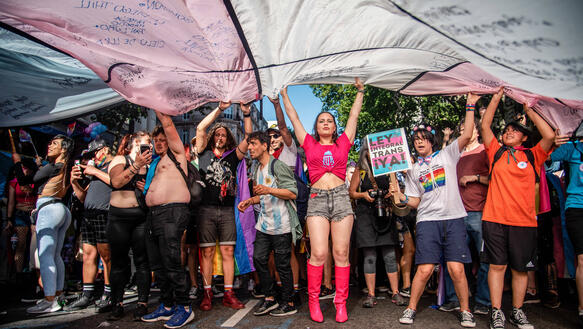 Mehrere Personen stehen auf einer Straße nebeneinander und breiten über ihren Köpfen eine große Transgender-Pride-Fahne aus.