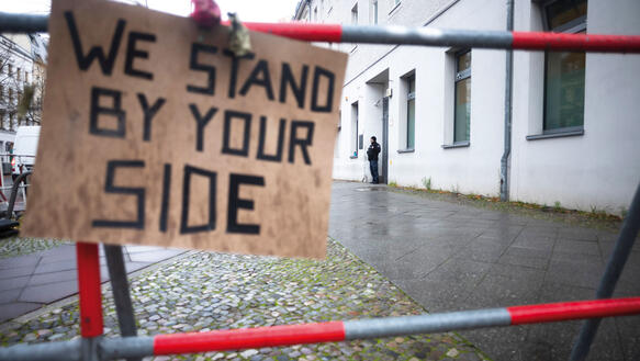 Eine Baustellenabsperrung in einer Berliner Straße, daran befestigt ein Schild auf dem per Hand geschrieben steht "We stand by your side".