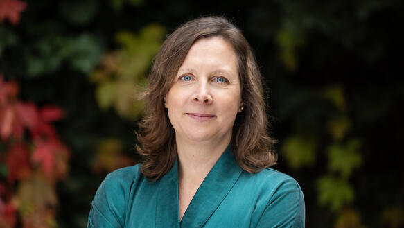 Eine Frau mit schulterlangem Haar steht in der Natur und verschränkt ihre Arme vor dem Oberkörper.