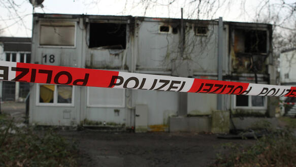 Wohncontainer, übereinandergestapelt, zum Teil ausgebrannt, abgesperrt mit einem Polizeiband.
