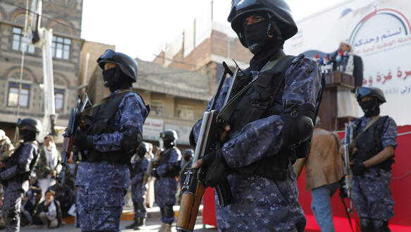 Das Foto zeigt mit Sturmgewehren bewaffnete Maskierte, die Helm und Uniform tragen und am Rande einer Demonstration Wache stehen.