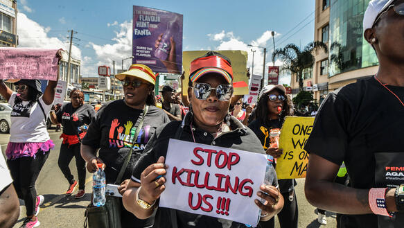 Männer und Frauen bei einer Demonstration auf einer Straße, sie halten Schilder hoch, auf einem steht "Stop Killing Us"