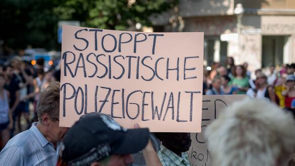 Das Bild zeigt mehrere Menschen, und ein Protestplakat auf dem steht "Stoppt rassistische Polizeigewalt"
