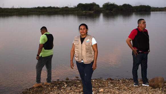 Das Bild zeigt drei Personen, die am Ufer eines Flusses stehen