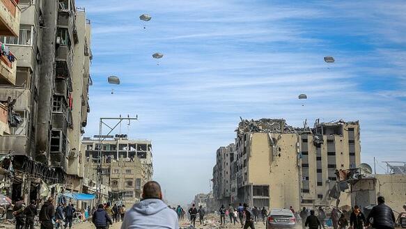 Das Bild zeigt im Vordergrund Menschen inmitten zerstörter Häuser, im Hintergrund am Himmel erkennt man mehrere Fallschirme
