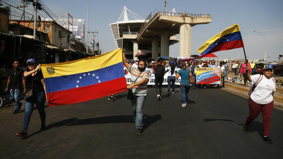 Das Bild zeigt viele Menschen auf einer breiten Straße, manchen halten die Nationalflagge Venezuelas in der Hand