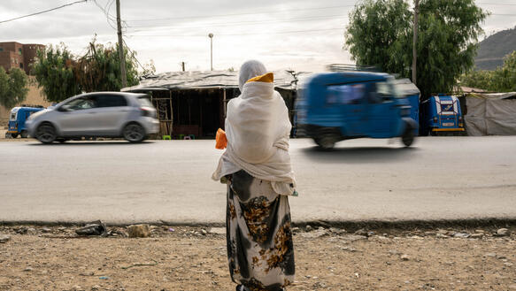 Das Foto zeigt eine Frau am Rande einer Straße, auf der Autos fahren. Sie trägt ein Gewand und ein Kopftuch und trägt ein Kind auf ihrem Rücken.