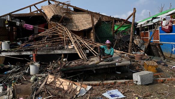 Das Bild zeigt eine Person, wie sie vor einem komplett zerstörten Haus sitzt