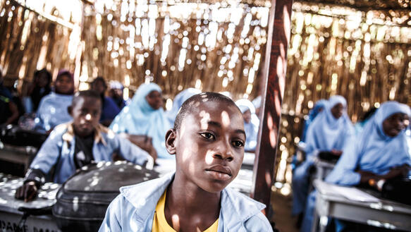 Ein Junge sitzt in einer Schule in Niger, einem Gebäude aus Holzzweigen, durch dessen Wände Sonne fällt, hinter ihm sitzen andere Schüler*innen, die Mädchen tragen Kopftuch.