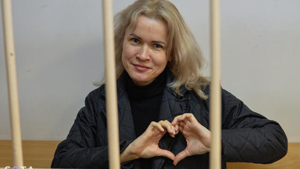 Das Bild zeigt das Porträtfoto einer Frau hinter Gitterstäben. Sie trägt Handschellen und formt ihre Hände zu einem Herzen und lächelt in die Kamera.