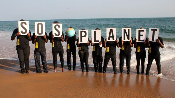 Das Foto zeigt zehn Personen, die jeweils Schilder mit Buchstaben vor ihre Gesichter halten. Dies ergibt den Schriftzug: "SOS Planet". Die vierte Person von rechts hält einen Globus vor ihr Gesicht.