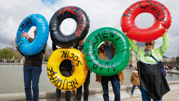 Menschen demonstrieren, sie halten Schwimmringe in den Händen, die an Olympische Ringe erinnern und beschriftet sind.