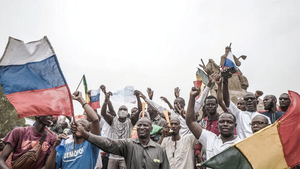 Menschen in Mali stehen mit wehenden Fahnen auf einer Straße und protestieren, viele heben die Hände, manche ballen sie zu einer Faust.