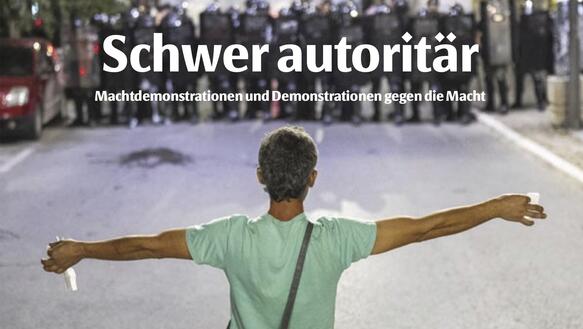 Ein Mensch steht auf einer Straße, er trägt T-Shirt, Jeans und eine Umhängetasche, er breitet die Arme aus, ihm gegenüber steht ein Block aus Sicherheitskräften mit Helmen und Schildern.