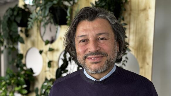 Porträtfoto von Taner Kılıç, aufgenommen vor einer mit Pflanzen geschmückten Wand. Er lächelt in die Kamera.