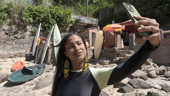 Eine junge indigene Frau mit langen Haaren in einem Schwimmhemd an einem Strand macht ein Selfie, sie trägt Ohrringe, hinter ihr erhebt sich eine Böschung vom Strand aus.