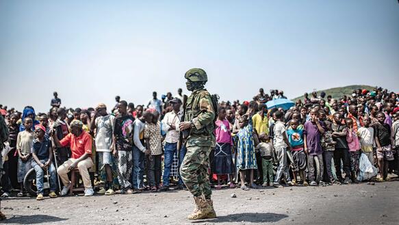 Ein Soldat patroulliert vor einer großen Menschenmenge; die Menschen drängen sich, Kinder, Männer, Frauen, Alte.