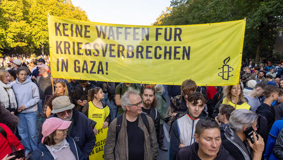 Das Foto zeigt mehrere Personen in einer riesigen Menschenmenge, die ein Banner hochhalten mit Amnesty-Logo und dem Schriftzug "Keine Waffen für Kriegsverbrechen in Gaza!"