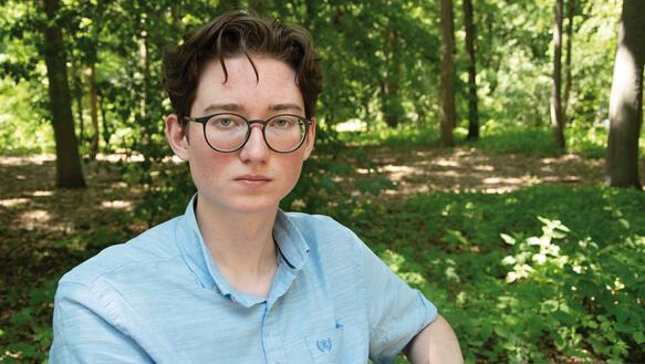 Ein junger Mann mit Brille trägt ein Hemd und sitzt draußen vor ein paar Bäumen.
