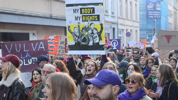 Das Foto zeigt einen Demonstrationszug auf der Straße in einer Stadt. Eine Teilnehmerin hält ein Schild hoch, auf dem steht: "My body, my rights!"