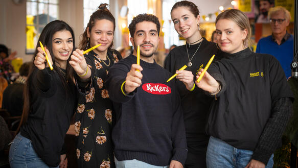Vier junge Frauen und ein junger Mann stehen nebeneinander und halten lächelnd Stifte in die Kamera.