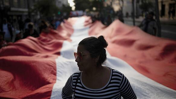 Eine peruanische Frau mit Sonnenbrille, die Haare zusammengebunden, trägt ein getreiftes Top, hinter ihr auf einer Straße hält eine Menschenmenge eine große Stoffbahn aufgespannt.