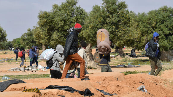 Mehrere Menschen mit Gepäck, Bündel stehen auf einem Feld, sie verlassen das Gelände