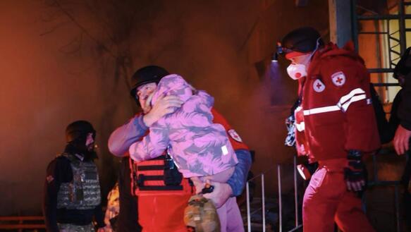 Rettungskräfte tragen ein kleines Kind aus einem Haus. Im Hintergrund lodert Feuer.