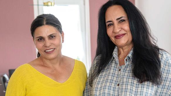 Zwei Frauen mittleren Alters stehen in einem Zimmer, beide haben ihre Händen in ihre Hosentaschen gesteckt.