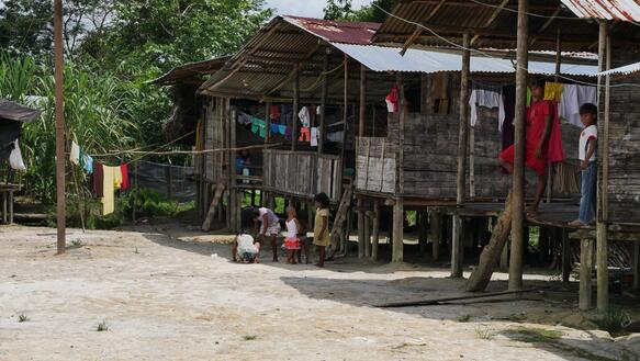 Das Foto zeigt mehrere Holzhütten, errichtet auf niedrigen Stelzen. Auf den Veranden stehen Menschen.