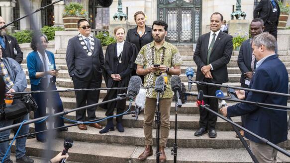 Ein junger Mann steht auf einer Treppe, vor ihm Mikrofone, er hält ein Handy in der Hand und spricht, um ihn herum stehen Männer und Frauen in formeller Kleidung.