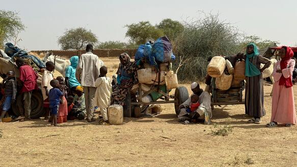Das Foto zeigt mehrere Frauen, Männer und Kinder neben vollgepackten Karren stehen oder neben ihnen auf dem kargen Boden sitzend.
