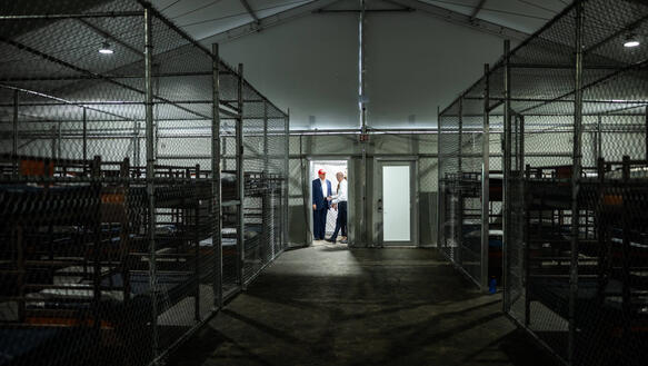 Das Foto zeigt ein großes Zelt, in dem mehrere Käfige mit Betten aufgestellt wurden. Weit hinten am Ende des Zeltes steht Donald Trump im Eingang mit einer weiteren Person.