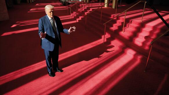 Donald Trump trägt Anzug und Krawatte und steht in einem Gebäude vor einer sechstufigen Treppe mit Messinggeländer.